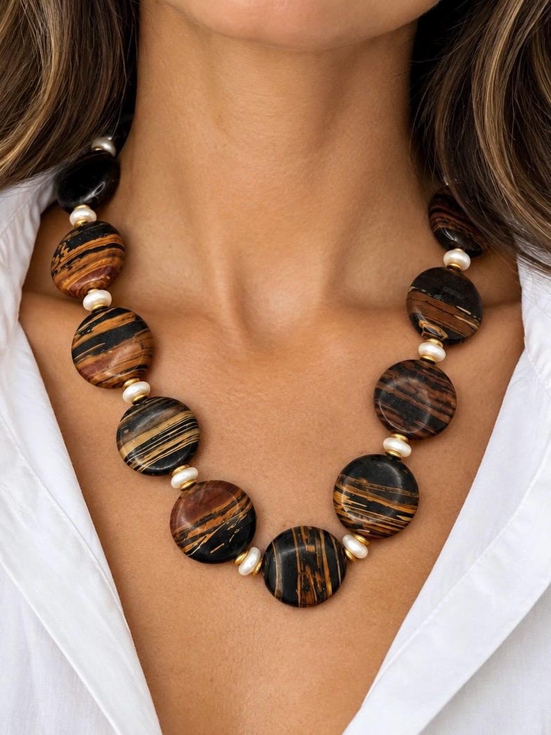 The Sutton necklace, with large round brown-and-black striped beads, small creamy white pearls and gold-tone accents, worn with a white open-collar shirt.