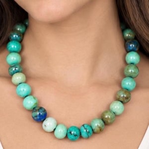 Op de afbeelding: Een kralenketting met ronde, gepolijste stenen in verschillende tinten turkoois, blauwgroen en diepblauw. De kralen vertonen natuurlijke marmering en kleurvariaties en zijn aan elkaar geregen tot een doorlopende cirkelvormige streng.