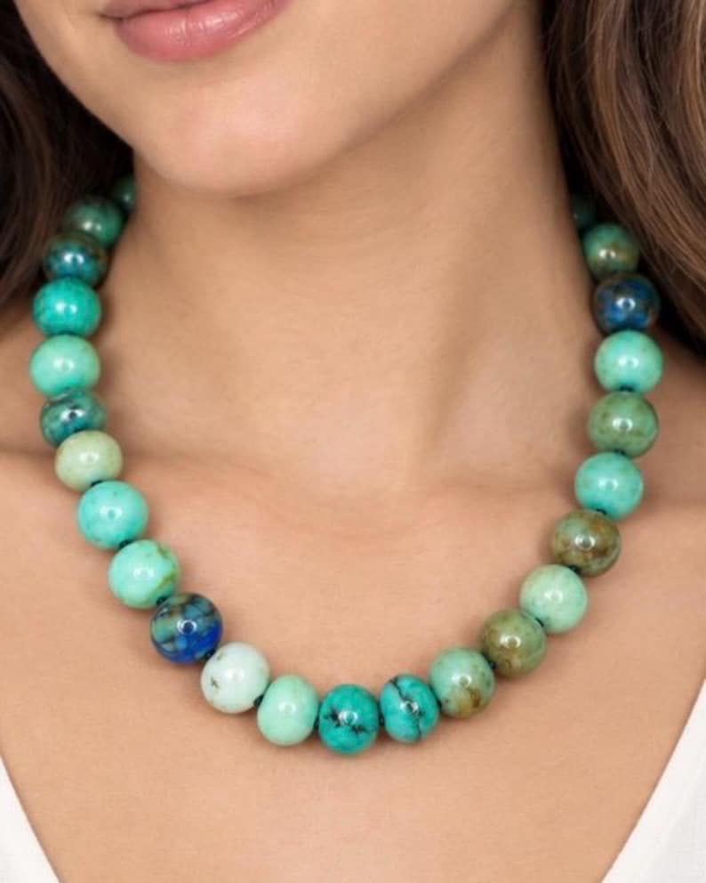 Op de afbeelding: Een kralenketting met ronde, gepolijste stenen in verschillende tinten turkoois, blauwgroen en diepblauw. De kralen vertonen natuurlijke marmering en kleurvariaties en zijn aan elkaar geregen tot een doorlopende cirkelvormige streng.