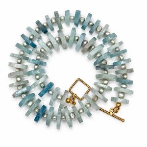 Peut inclure: Un collier compos&eacute; d'une alternance de perles de pierre rectangulaires bleu clair et de petites perles rondes grises. Le bijou est ferm&eacute; par un fermoir &agrave; bascule dor&eacute;, form&eacute; d'une boucle carr&eacute;e et d'une barre en T, dispos&eacute; en cercle.