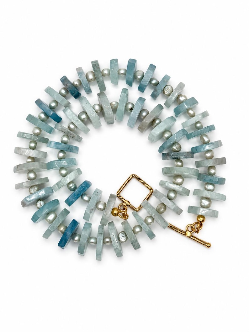 Peut inclure: Un collier compos&eacute; d'une alternance de perles de pierre rectangulaires bleu clair et de petites perles rondes grises. Le bijou est ferm&eacute; par un fermoir &agrave; bascule dor&eacute;, form&eacute; d'une boucle carr&eacute;e et d'une barre en T, dispos&eacute; en cercle.