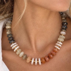 May include: A beaded necklace featuring a gradient of smooth, round, and disc-shaped beads in shades of grey, white, cream, and orange. The beads are separated by small, polished gold-tone metal spacers, creating a structured, multi-coloured design.