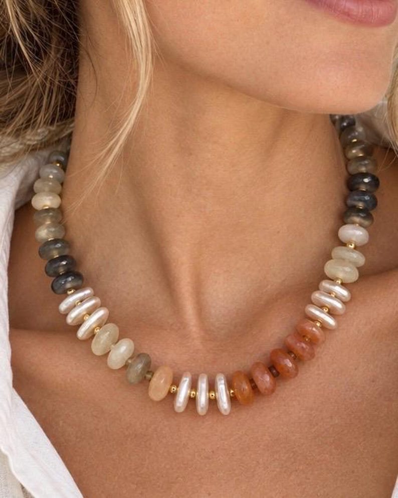 May include: A beaded necklace featuring a gradient of smooth, round, and disc-shaped beads in shades of grey, white, cream, and orange. The beads are separated by small, polished gold-tone metal spacers, creating a structured, multi-coloured design.