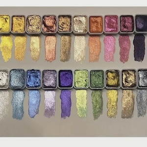 May include: A set of twenty-eight rectangular watercolor pans arranged in two rows, each containing metallic pigment. Below each pan is a corresponding vertical swatch of the paint, displaying a range of shimmering colors including gold, copper, pink, purple, blue, green, and silver tones.