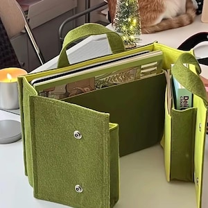 May include: An olive green felt organizer bag with two top handles, featuring multiple interior compartments and exterior pockets. The bag is secured with silver metal snap buttons and contains a book and a small box labeled Delicious.