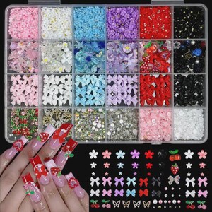 May include: A clear plastic storage container filled with twenty-four compartments of assorted nail art charms, including miniature flowers, bows, cherries, and rhinestones, alongside a display of matching nail art designs on fingernails.
