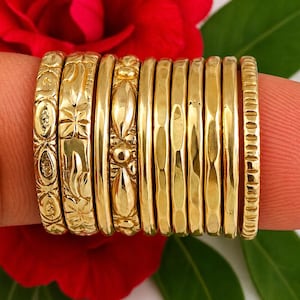 May include: A set of ten gold-toned rings arranged in a row. The rings feature various textures and patterns, including floral-inspired engravings, smooth surfaces, hammered finishes, and a band with vertical indentations along the outer edge.