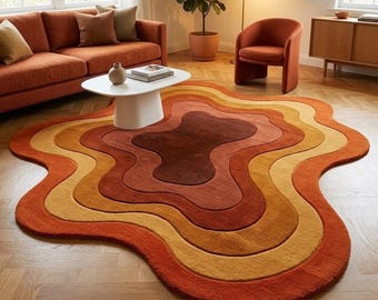 May include: An irregular, organic-shaped area rug featuring concentric, wavy layers in shades of dark brown, rust orange, burnt orange, and golden yellow, creating a topographical, multi-layered effect on a wooden floor.