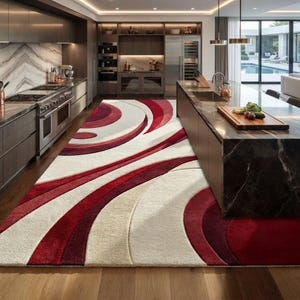 May include: A large rectangular area rug featuring a bold, abstract pattern of sweeping, concentric curves in shades of deep crimson, bright red, and cream. The rug has a thick, plush texture and covers the floor between a kitchen island and cabinets.
