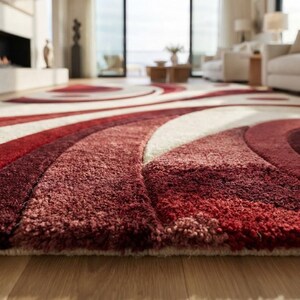 May include: A plush area rug featuring a high-pile texture with a bold, abstract pattern of sweeping curves and circular shapes. The design incorporates deep shades of burgundy, vibrant red, and soft cream, creating a layered visual effect.