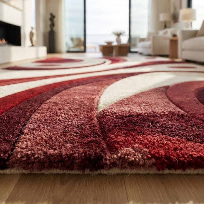 May include: A plush area rug featuring a high-pile texture with a bold, abstract pattern of sweeping curves and circular shapes. The design incorporates deep shades of burgundy, vibrant red, and soft cream, creating a layered visual effect.