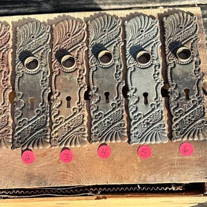 May include: Six rectangular metal door plates with ornate embossed scrollwork and floral patterns. Each plate features a circular opening for a knob and a smaller keyhole below. The plates are numbered one through six with small pink circular stickers.