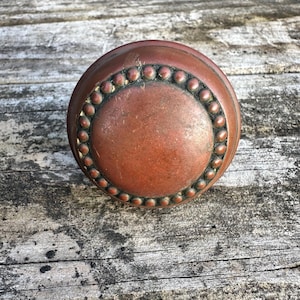 May include: A round, copper-toned metal door knob featuring a smooth, slightly weathered central surface surrounded by a decorative border of small, raised circular beads. The object has a warm, metallic finish with visible signs of age.
