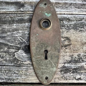 May include: An oval-shaped brass door plate featuring a weathered, tarnished finish with visible green oxidation. The plate includes a central circular opening for a door knob, a lower keyhole, and two small screw holes at the top and bottom.