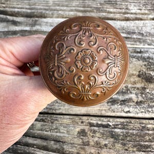May include: A round, copper-toned metal door knob featuring an embossed, symmetrical floral and scrollwork pattern across its surface. The design centers on a small flower motif surrounded by intricate, swirling decorative elements in relief.