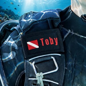 以下が含まれることがあります： 赤色の文字でTobyと刺繍され、赤と白のダイバーダウンフラッグが描かれた長方形のパッチが付いた黒いスキューバダイビング用BCDストラップ。ストラップには銀色の金属製Dリングと灰色のプラスチック製金具が取り付けられています。