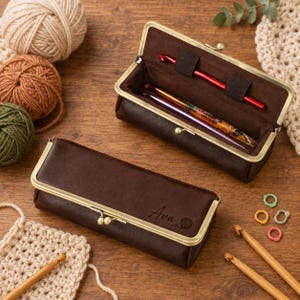 May include: Two dark brown rectangular cases with brass-toned metal frames and kiss-lock closures. One case is closed and features the name Ava engraved with a yarn ball icon. The other case is open, revealing crochet hooks inside.