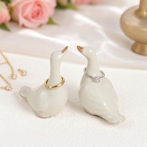 May include: Two off-white ceramic goose-shaped ring holders. One goose holds a plain gold band around its neck, while the other holds a silver ring with a single clear stone setting. The figures have smooth, glossy finishes and orange beaks.