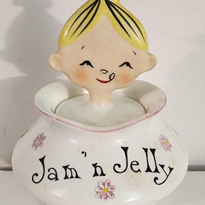 May include: A white ceramic jar with a lid featuring a sculpted figure of a child with yellow hair and a pink bow. The jar is decorated with small pink and yellow flowers and the black text Jam 'n Jelly printed on the front of the rounded body.
