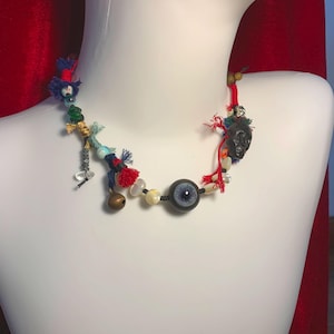 May include: A necklace featuring a central blue eye bead, a small brass bell, a dark carved skull bead, and various colorful glass beads interspersed with red and blue fabric tassels on a dark cord, displayed on a white mannequin neck.