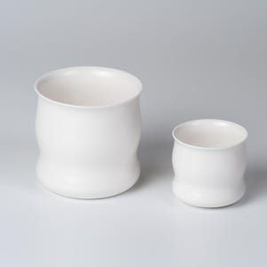 May include: Two white ceramic vessels with a smooth, matte finish and a curved, rounded silhouette. The larger vessel is positioned on the left, while a smaller, matching vessel sits to its right, both featuring open tops.