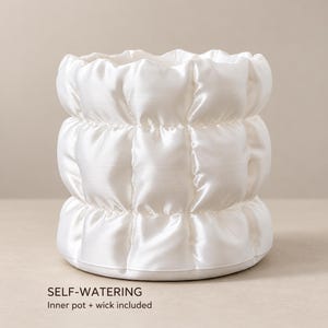 May include: A white cylindrical self-watering planter with a quilted, puffy texture. The exterior features a grid-like pattern of soft, rounded sections. Text at the bottom reads: SELF-WATERING, Inner pot + wick included.
