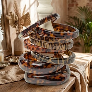 May include: A stack of five circular bangle bracelets featuring a glossy brown and black tortoiseshell pattern. The bracelets are made of a smooth, translucent resin material and are displayed stacked vertically on a white stand.