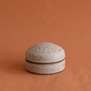 Op de afbeelding: Een kleine, ronde keramische houder met deksel in een gespikkelde, gebroken witte matte afwerking. De houder heeft een koepelvormig deksel dat naadloos aansluit op de basis, wat zorgt voor een eenvoudig cilindrisch silhouet met een bruine naad.