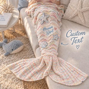 Puede incluir: Una manta de cola de sirena tejida en hilo multicolor pastel con una textura ondulada. La manta está decorada con aplicaciones tridimensionales de estrellas de mar y conchas marinas e incluye el texto bordado Ocean Girl con un pequeño corazón.