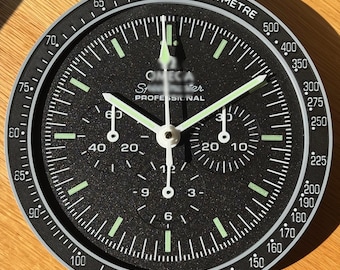 May include: A round black wall clock designed to resemble a chronograph watch face. It features a tachymeter scale on the outer bezel, three sub-dials, luminous hour markers, and white hands. The text OMEGA Speedmaster PROFESSIONAL is printed on the dial.
