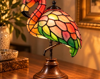 Op de afbeelding: Een glas-in-lood tafellamp in de vorm van een flamingo met een gebogen nek en een lichaam met panelen van roze, oranje, geel en groen glas, verbonden door donkere metalen lijnen, op een ronde, donkerbruine houten voet.