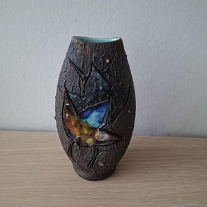 Puede incluir: Un jarrón de cerámica con un exterior de color marrón oscuro, fuertemente texturizado y similar a la corteza. La superficie presenta un área central lisa y hundida, decorada con un esmalte brillante en tonos azul, verde y ámbar.