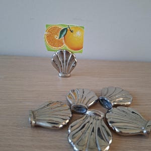 Könnte beinhalten: Ein Set aus silberfarbenen, muschelförmigen Platzkartenhaltern aus Metall. Ein Halter steht aufrecht und enthält eine kleine rechteckige Karte mit der Grafik einer aufgeschnittenen Orange und einer ganzen Orange mit grünem Blatt. Fünf weitere Muscheln liegen flach.