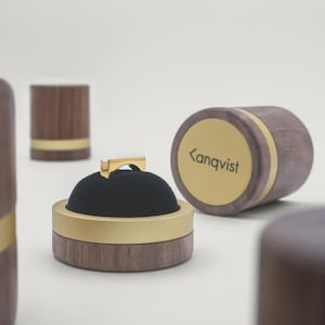Op de afbeelding: Een gouden ring met een witte bovenkant rust op een zwart fluwelen kussentje in een ronde houten doos met een gouden rand. Een bijpassend houten deksel met een gouden ronde plaat met de tekst Kanqvist ligt ernaast op een ondergrond.