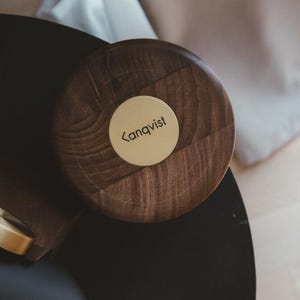 Op de afbeelding: Een cirkelvormig donkerbruin houten deksel met een zichtbare houtnerf. In het midden van het deksel bevindt zich een ronde, lichtgekleurde metalen schijf met de zwarte tekst "kanqvist" voorafgegaan door een kleiner-dan-teken.
