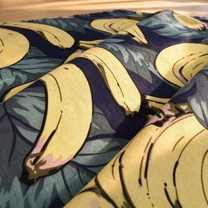 Könnte beinhalten: Ein gefalteter Stoff mit einem sich wiederholenden Muster aus gelben Bananen und gedämpften grünen Blättern auf dunkelblauem Hintergrund. Das Textil weist eine sichtbare Webstruktur auf und ist drapiert, um Tiefe zu zeigen.