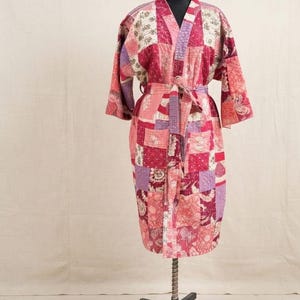 Op de afbeelding: Een knielange kamerjas met een patchworkontwerp in diverse tinten roze, rood en paars. Het kledingstuk heeft lange mouwen, een V-hals en een bijpassende stoffen ceintuur in de taille. De stof is voorzien van een doorgestikte textuur met bloemen.