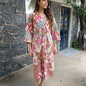 Puede incluir: Una bata acolchada hasta la rodilla con un diseño de patchwork que presenta varios patrones florales y geométricos en tonos rosa, azul, marrón y crema. La bata tiene mangas largas, cuello en V y un cinturón de tela a juego en la cintura.