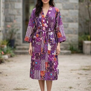 Könnte beinhalten: Ein knielanger Morgenmantel mit Patchwork-Design in Lila-, Orange- und Weißtönen. Das Kleidungsstück hat lange Ärmel, einen V-Ausschnitt und einen passenden Bindegürtel aus Stoff in der Taille. Der Stoff zeigt verschiedene Blumen- und Geometriemuster.