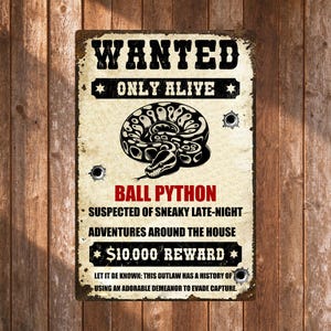 May include: A rectangular metal sign with a distressed cream background featuring the text WANTED ONLY ALIVE in black. A black and white illustration of a coiled ball python is centered. Red text reads BALL PYTHON above black text details.