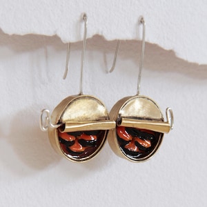 May include: A pair of drop earrings featuring circular gold-toned metal casings that resemble open tin cans. Each earring has a partially peeled gold lid and contains a textured interior filled with glossy, alternating black and orange resin.