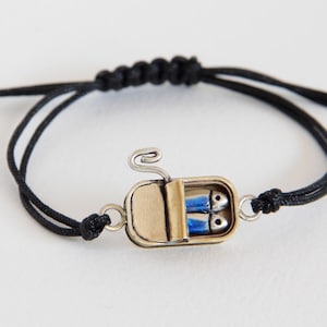Può includere: Bracciale originale con un ciondolo a forma di scatola di sardine contenente due pesci blu. Il ciondolo è argentato e dorato, montato su un braccialetto di cordino nero. Il bracciale è regolabile.