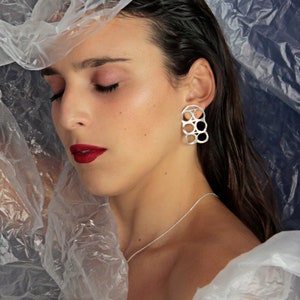 Puede incluir: Una mujer con pendientes geométricos plateados con un diseño circular. Los pendientes son grandes y cuelgan de sus orejas. Lleva una camisa blanca y tiene el pelo recogido hacia atrás.