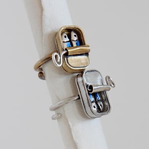 Peut inclure: Deux bagues en forme de boîte de sardines. L'une est dorée, l'autre argentée. Chaque bague représente une boîte de sardines miniature avec deux poissons bleus à l'intérieur. Les boîtes ont un couvercle à charnière.
