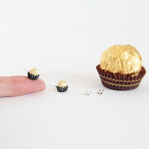 May include: A pair of gold and brown chocolate-shaped earrings with silver posts. The earrings are a miniature version of a popular chocolate candy.