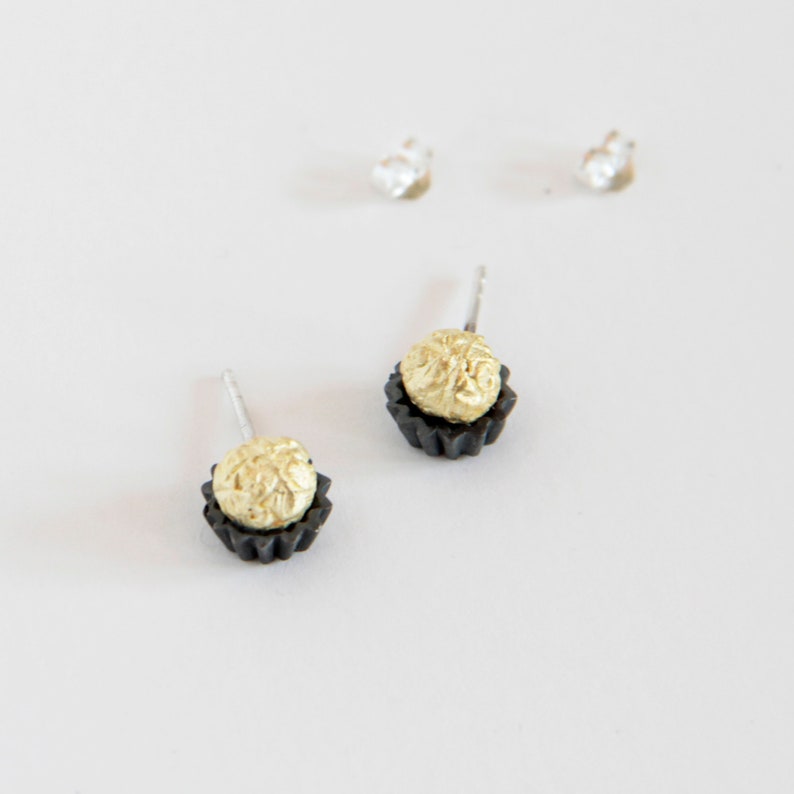 May include: A pair of gold and black stud earrings. The earrings are shaped like small cupcakes with a gold center and a black outer rim.