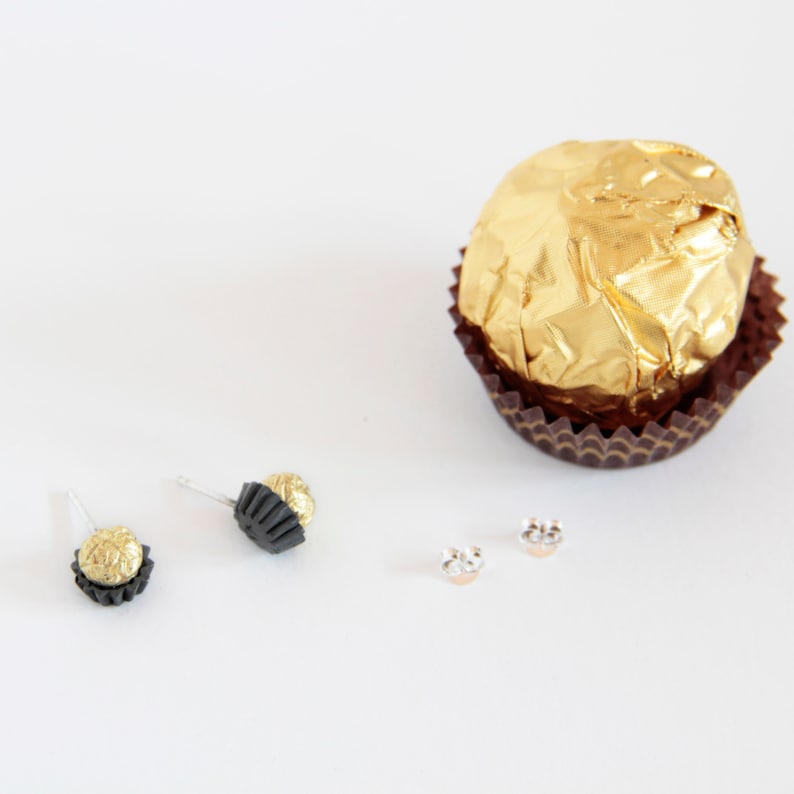 May include: A pair of gold and black earrings shaped like chocolate truffles. The earrings are on a white surface next to a single chocolate truffle wrapped in gold foil.