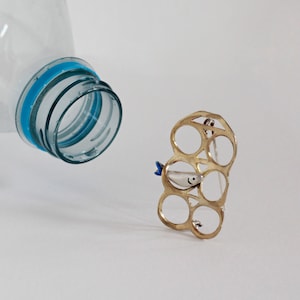 Puede incluir: Un pin de metal de color dorado con forma de anillo de seis latas con un pequeño amuleto de pez blanco adjunto. El pez tiene una cola azul y un ojo negro.