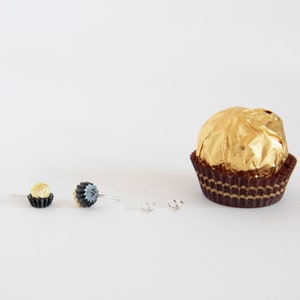 May include: A pair of earrings featuring a chocolate truffle design. The earrings are made of black polymer clay and have a gold foil finish. The earrings are shown next to a single chocolate truffle wrapped in gold foil.