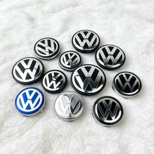 May include: A collection of eleven circular Volkswagen wheel centre caps. Ten caps feature a black and chrome finish with the iconic VW logo, while one cap displays a vibrant blue and chrome finish. Each piece is round with a smooth, reflective surface.
